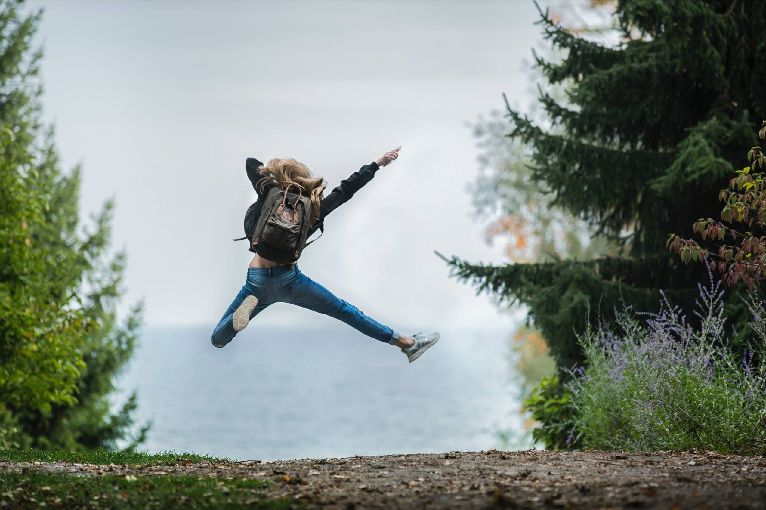 Omslagsbild till undersökningen om ungas psykiska hälsa, en ungdom med ryggsäck fotograferad bakifrån som hoppar högt på en grusväg kantad av stora granar.